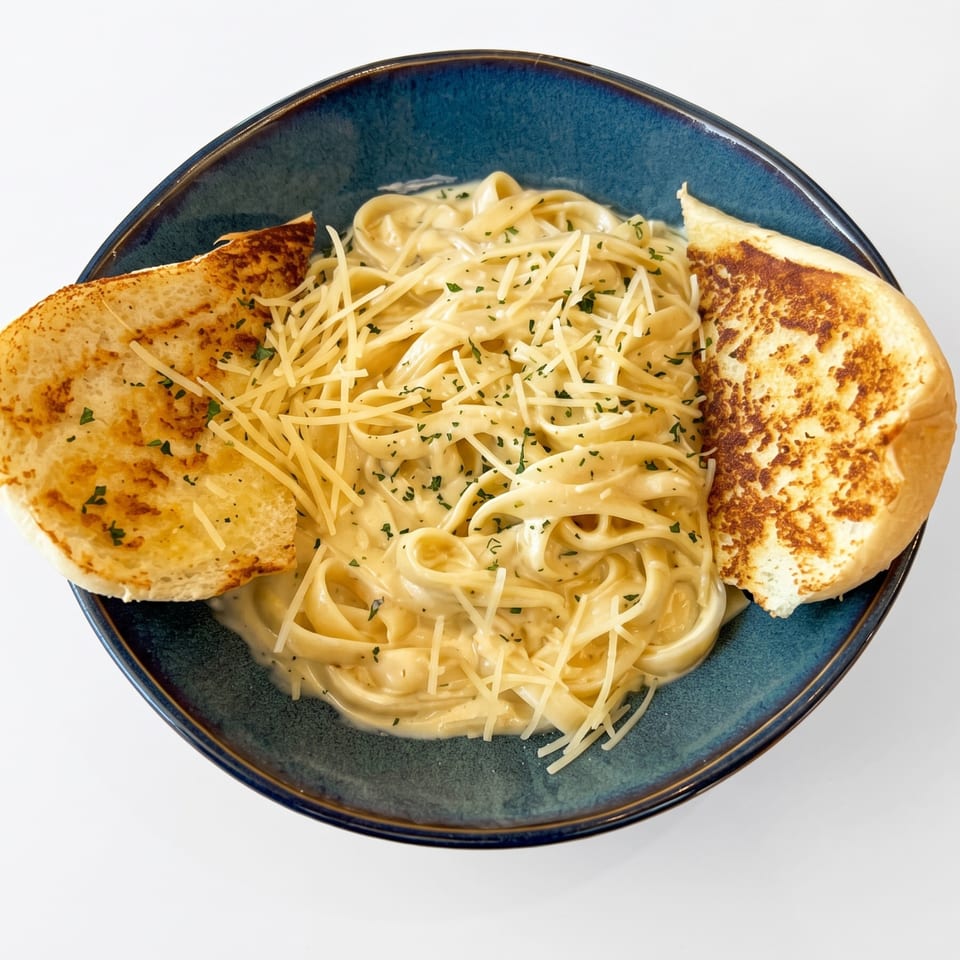 Creamy fettuccine alfredo pasta topped with cheese and parsley, served with garlic bread in a blue bowl.