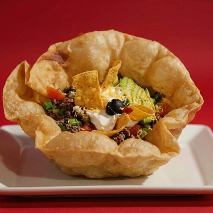 A taco salad in a crispy fried tortilla bowl, topped with ground beef, avocado, and sour cream.