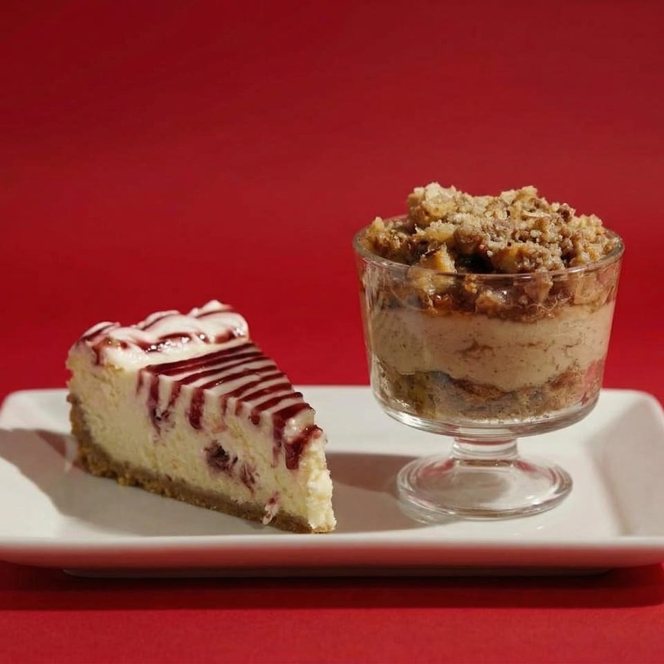 A slice of cheesecake with berry drizzle and a parfait with crumble topping on a white plate.