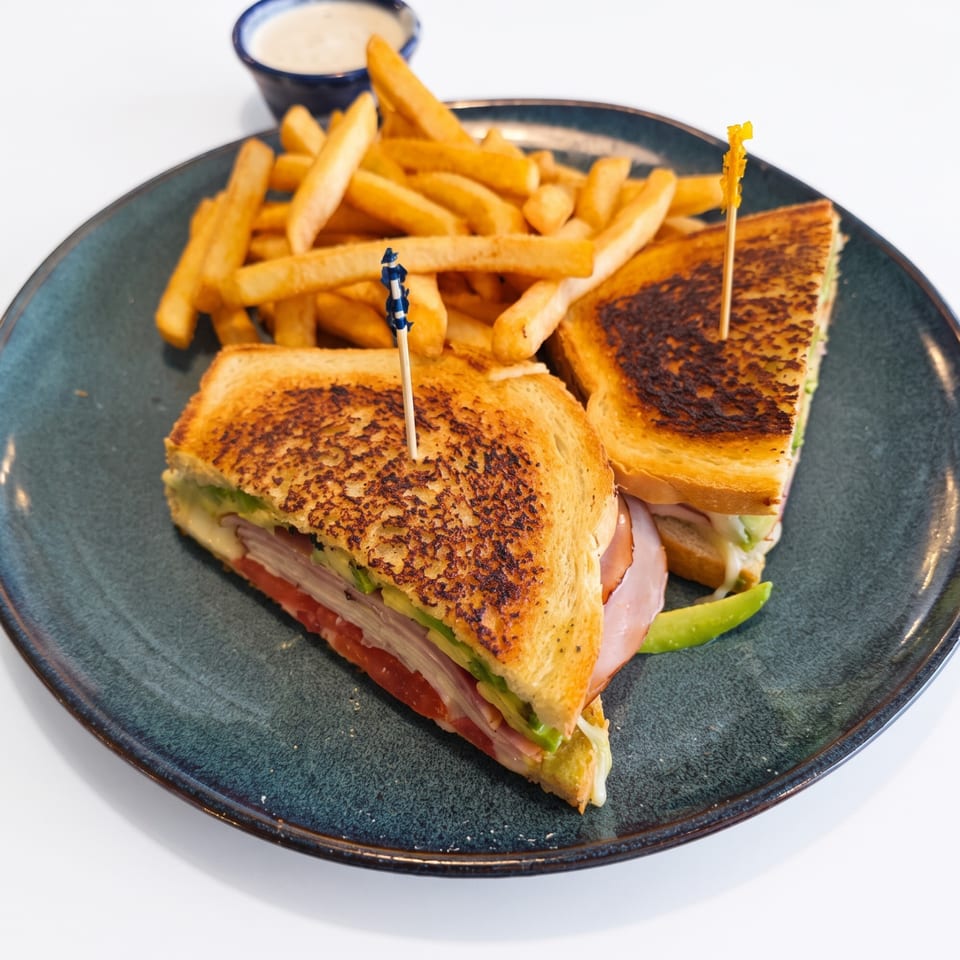Toasted sandwich with ham, avocado, and tomato, accompanied by golden french fries and dipping sauce.